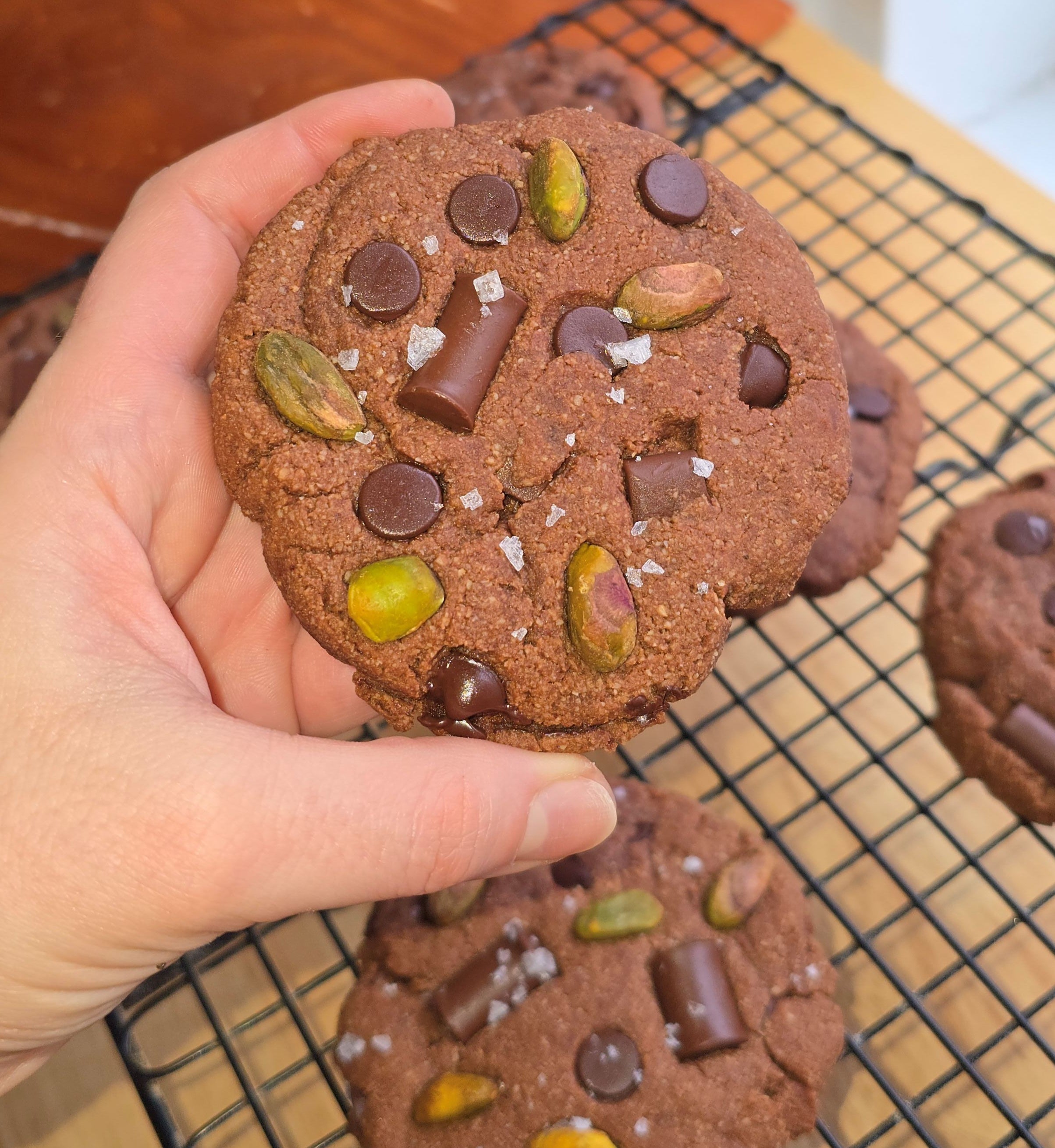 Hand holding a chocolate cookie with nuts and chocolate chips, more cookies on a cooling rack in the background.