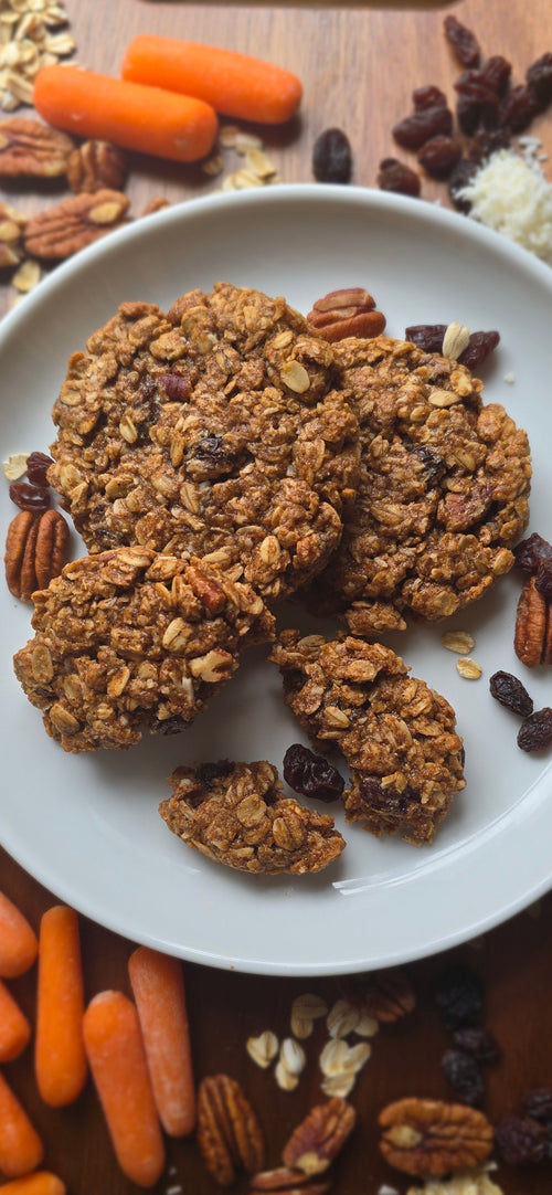Oatmeal cookies on a white plate with carrots, oats, and nuts on a wooden surface