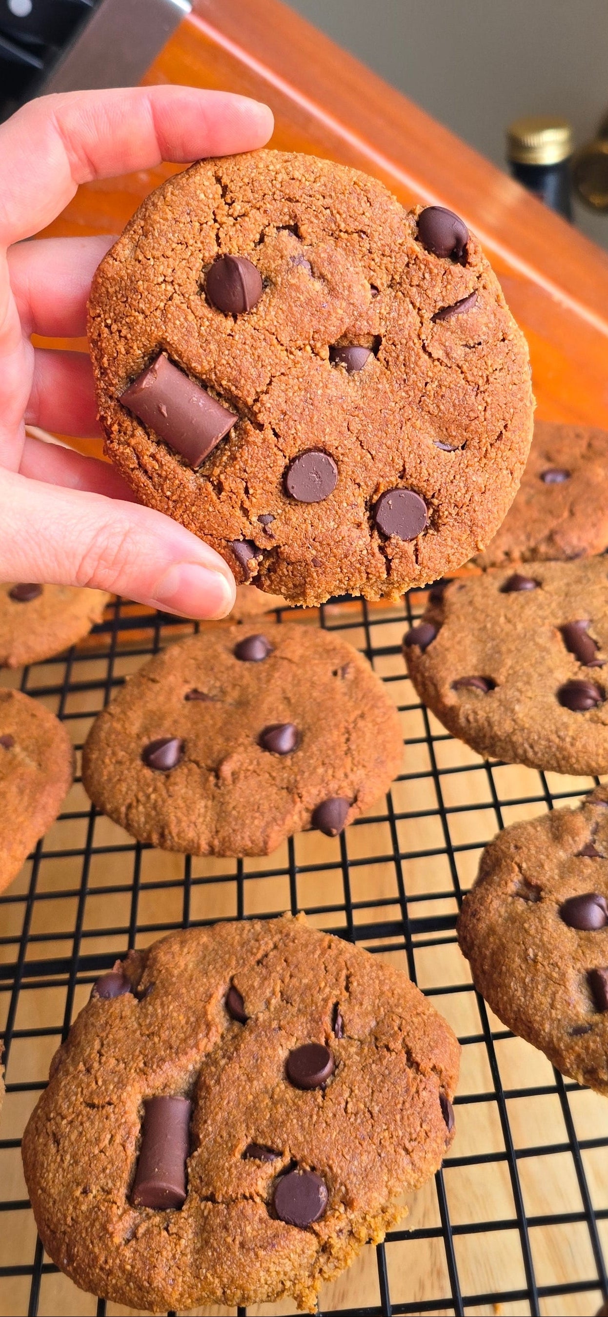 Hand holding a chocolate chip cookie with more cookies on a cooling rack in the background.